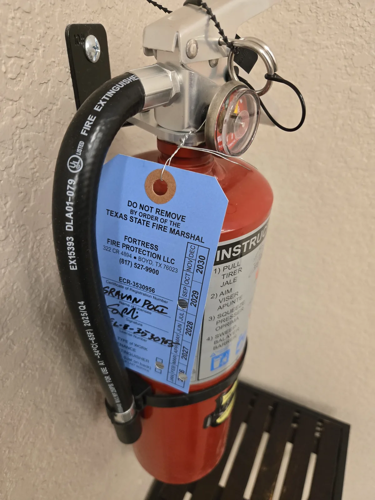 A signed and dated Fortress Fire Protection inspection tag on a fire extinguisher, listing the firm's Texas registration number